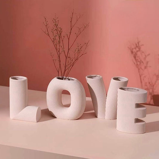 Decorative ceramic letters spelling 'LOVE' with a vase containing dried branches on a pink background.